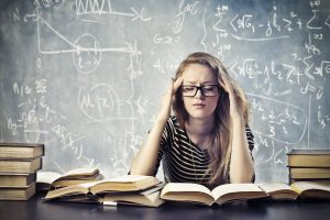 A young woman wearing glasses and a black and white striped shirt holds her temples as she is surrounded by text books.